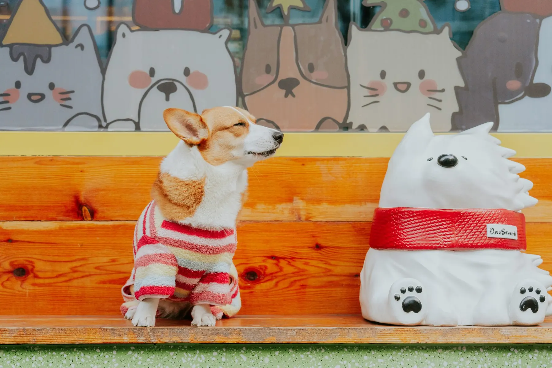 A corgi in a striped sweater beside a white dog statue, in front of a painted mural of pets
